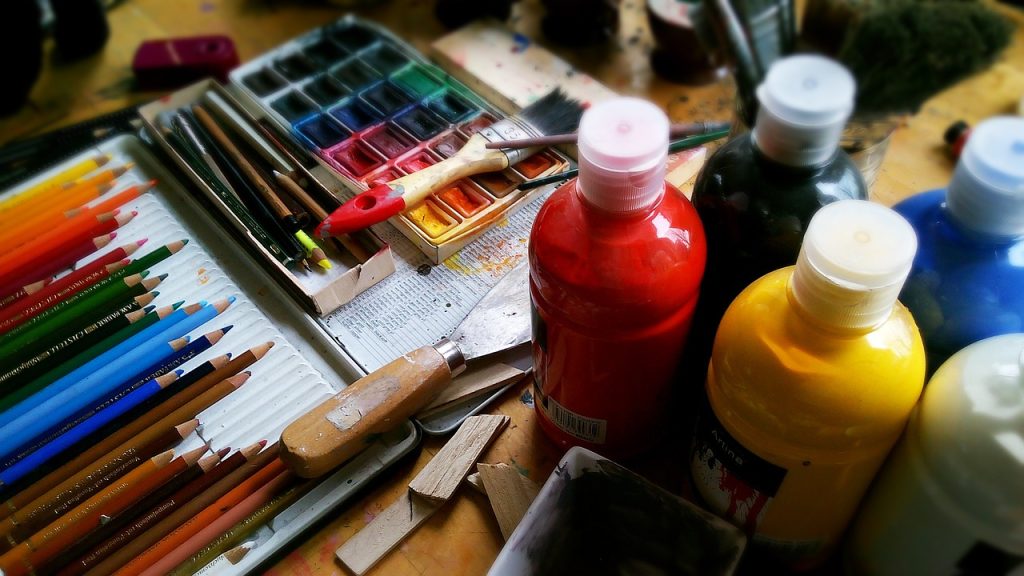 Ensemble de matériel d'artiste comprenant des crayons de couleur, une palette d'aquarelle, des pinceaux, des bouteilles de peinture acrylique et un couteau à peindre, disposés sur une table de travail.
