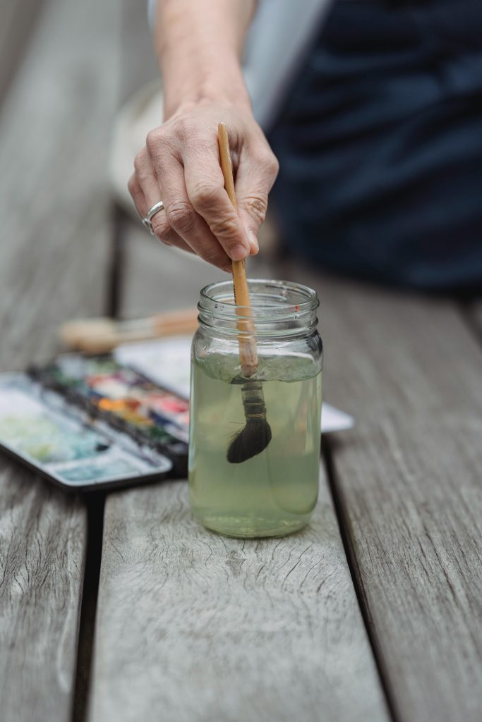Main d'une personne nettoyant un pinceau dans un pot en verre rempli d'eau teintée, avec une palette d'aquarelle posée sur une table en bois en arrière-plan.