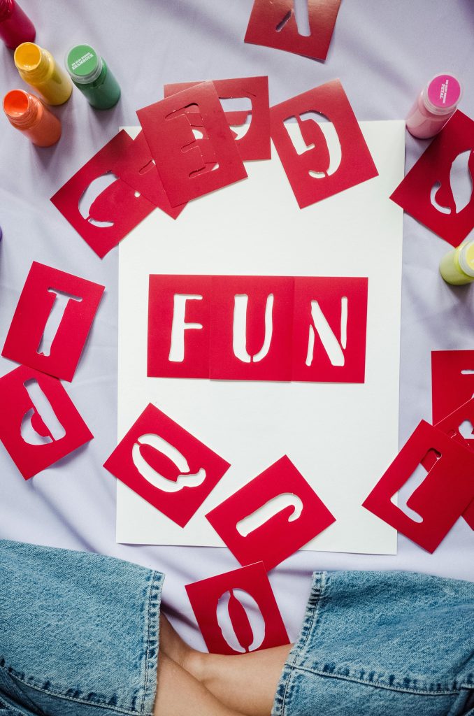 Pochoirs rouges en forme de lettres disposés autour d'une feuille blanche, avec le mot "FUN" écrit au centre. Des pots de peinture colorés et une personne en jean sont visibles en arrière-plan.